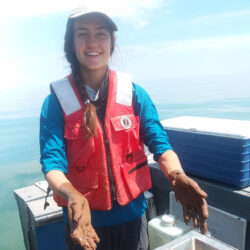 Audrey Huff is smiling at the camera while conducting aquatic research. She is wearing a baseball cap and a red life jacket. Her hands are covered in mud.