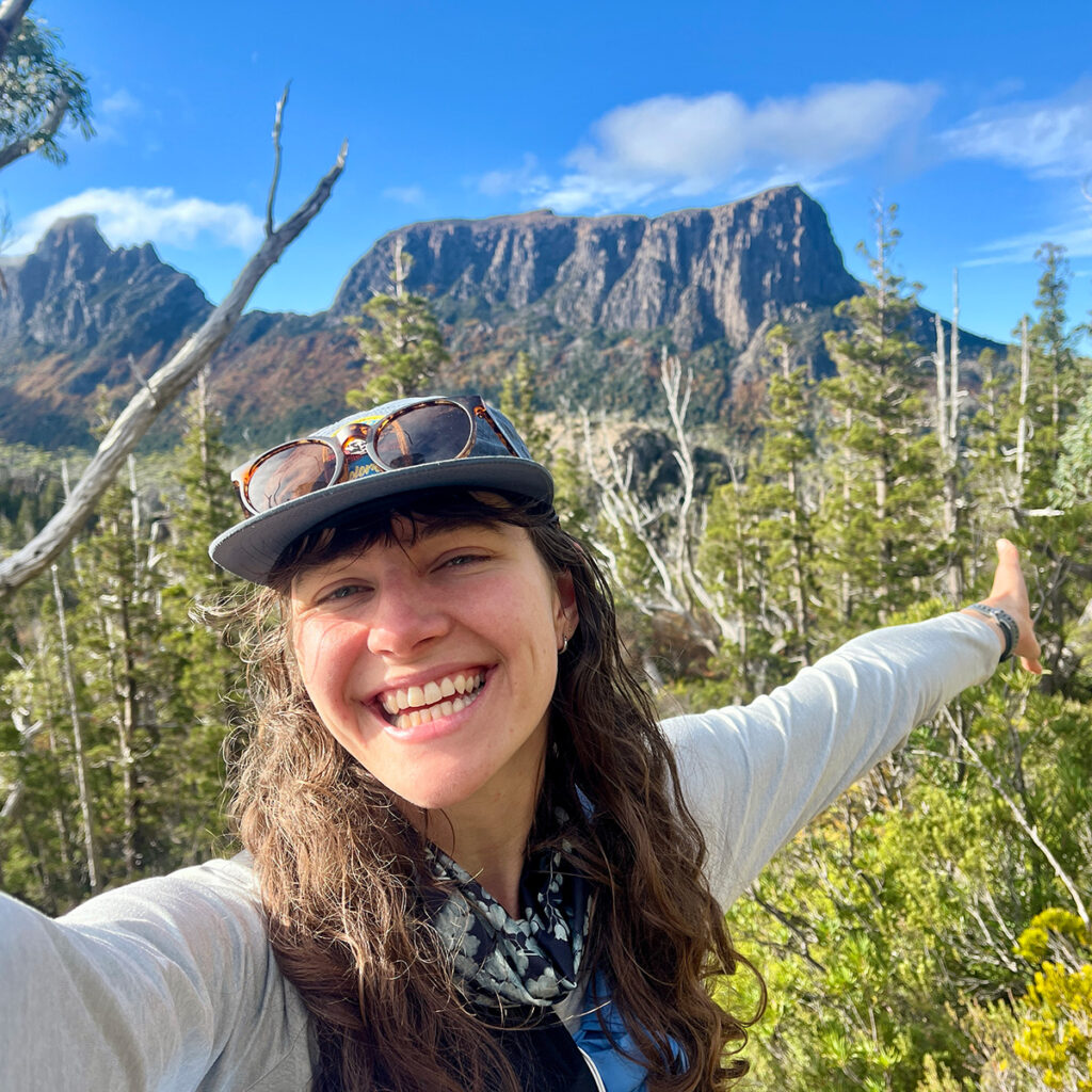 Aurelia is smiling at the camera, arms outstretched, with an evergreen forest and a mountain plateau in the background.