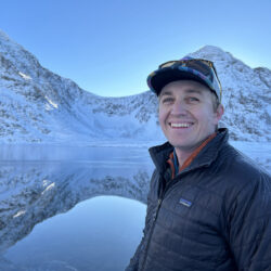 Dusty Reed smiling at the camera in a black puffy jacket, with a calm lake and snow-capped mountains behind him.
