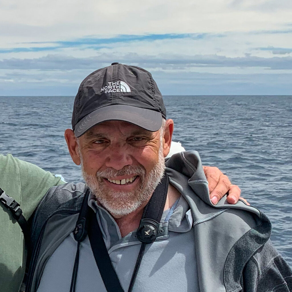 Researcher Marc Webber is smiling at the camera with his back to the ocean. He is wearing a black baseball cap, a grey hoodie, and a slate blue shirt. The straps of his binoculars are visible around his neck.