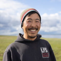 Photo of Ryan Choi. Ryan is smiling with a graminoid meadow and white clouds behind him.